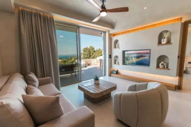 Μοντέρνο σαλόνι με άνετο καναπέ, πολυθρόνα, ξύλινο τραπεζάκι και τηλεόραση σε εντοιχισμένο τοίχο, με μεγάλα τζαμωτά που ανοίγουν σε βεράντα με θέα στη θάλασσα.