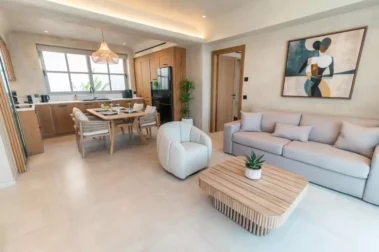 Ανοιχτή κουζίνα με ξύλινα ντουλάπια, τραπέζι φαγητού, σύγχρονο καθιστικό με καναπέ, πολυθρόνα και ξύλινο coffee table, διακοσμημένος με αφηρημένο έργο τέχνης.