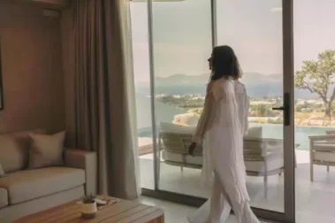 Γυναίκα με λευκά ρούχα στέκεται μπροστά σε τζαμαρία σαλονιού με ξύλινο τραπεζάκι και θέα θάλασσα και πισίνα.