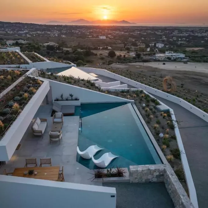 Πανοραμική άποψη βίλας με ιδιωτική πισίνα, εξωτερικό lounge και τραπέζι φαγητού, με φόντο το ηλιοβασίλεμα στο Αιγαίο.