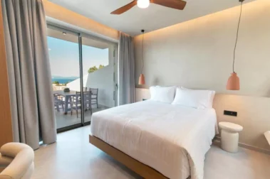 Φωτεινό δωμάτιο με μεγάλο κρεβάτι, ξύλινα έπιπλα και μπαλκονόπορτα που οδηγεί σε βεράντα με θέα.