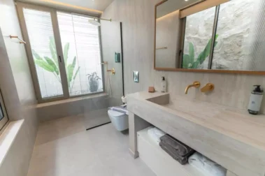 Minimal μπάνιο με walk-in ντουζιέρα, μεγάλα παράθυρα, εντοιχισμένο νεροχύτη με ξύλινο πάγκο και χρυσές λεπτομέρειες.