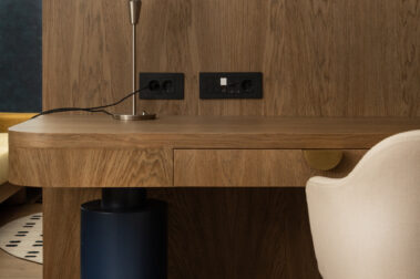 Ξύλινο γραφείο/πάγκος δωματίου ξενοδοχείου με μικρό μεταλλικό φωτιστικό, πρίζες στον τοίχο και λευκή καρέκλα στο πλάι