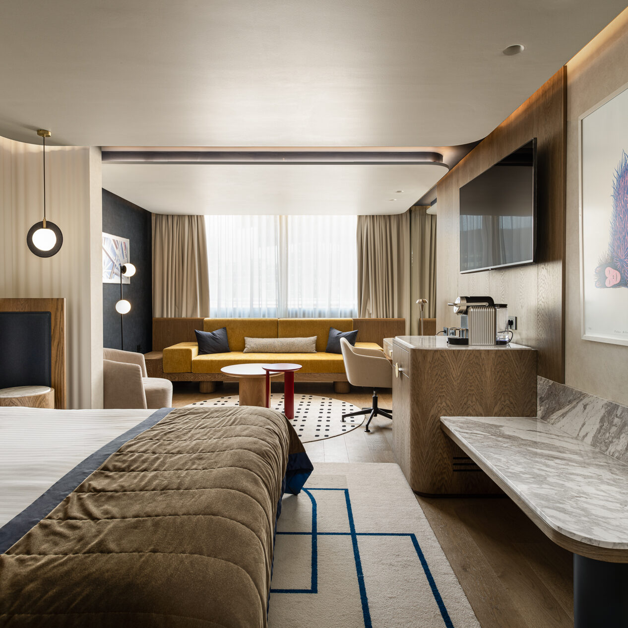 Ευρύ πλάνο σουίτας: κρεβάτι στο προσκήνιο, καθιστικό με κίτρινο καναπέ στο βάθος, ξύλινα έπιπλα και έργο τέχνης στον τοίχο.