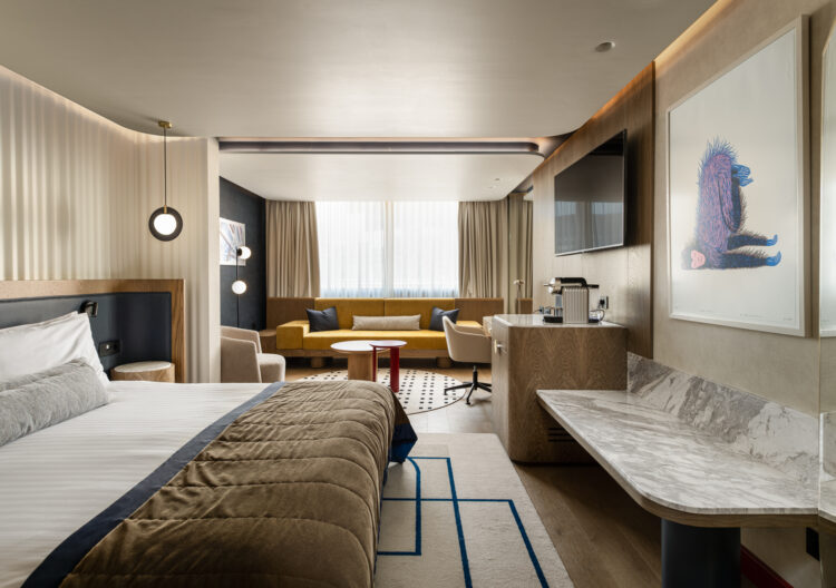 Ευρύ πλάνο σουίτας: κρεβάτι στο προσκήνιο, καθιστικό με κίτρινο καναπέ στο βάθος, ξύλινα έπιπλα και έργο τέχνης στον τοίχο.