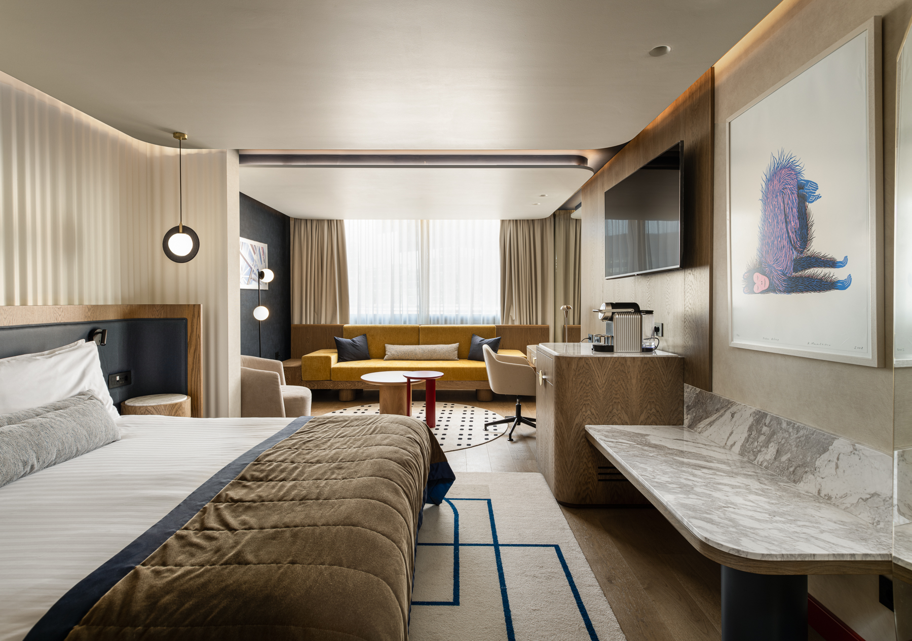 Ευρύ πλάνο σουίτας: κρεβάτι στο προσκήνιο, καθιστικό με κίτρινο καναπέ στο βάθος, ξύλινα έπιπλα και έργο τέχνης στον τοίχο.