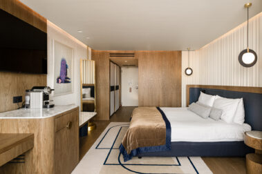 Πανοραμική άποψη δωματίου με κρεβάτι, ξύλινες επενδύσεις, πάγκο με καφετιέρα, χαλί με μπλε γεωμετρικές γραμμές και ζεστό κρυφό φωτισμό οροφής