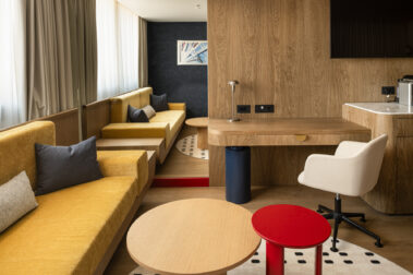 Καθιστικό χώρου με κίτρινους καναπέδες, στρογγυλά τραπεζάκια (ξύλινο και κόκκινο), χαλί με μαύρες τελείες και γραφείο στο βάθος.