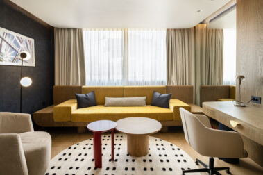 Μετωπικό πλάνο καθιστικού: κίτρινος καναπές μπροστά από μεγάλα παράθυρα με κουρτίνες, δύο στρογγυλά τραπεζάκια και γραφείο δεξιά