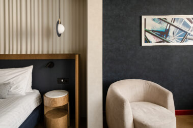 Γωνία δωματίου με μέρος κρεβατιού, στρογγυλό μαρμάρινο κομοδίνο, κρεμαστό φωτιστικό και μπεζ πολυθρόνα δίπλα σε σκούρο μπλε τοίχο.